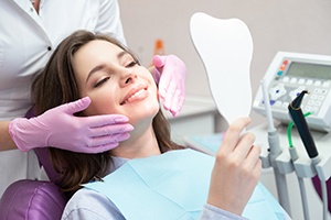 Dentist’s gloved hands touching cheeks of woman in dental chair smiling at tooth-shapred mirror